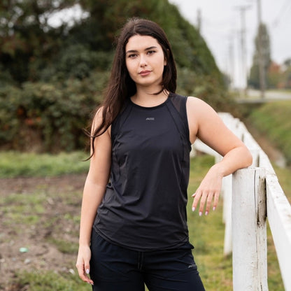Woman in a black sleeveless top and pants standing by a white fence with greenery in the background