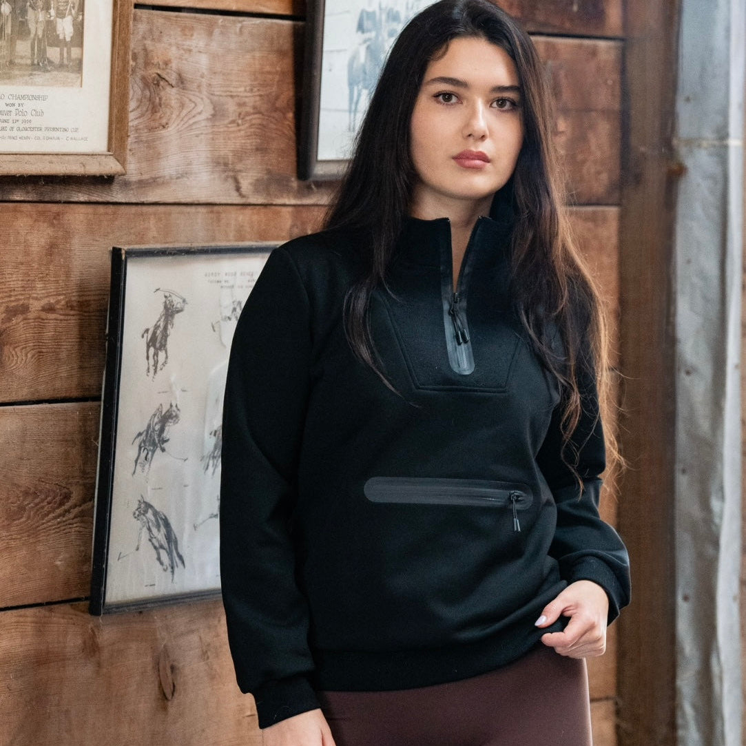Woman wearing a black pullover and brown leggings standing against a wooden wall with framed pictures.