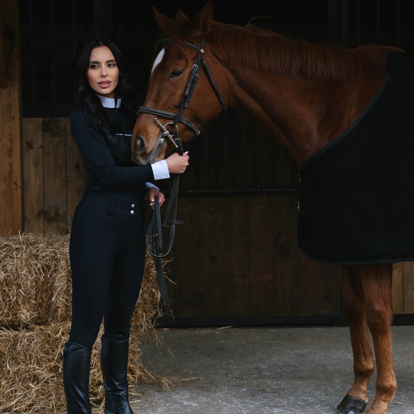 Woman in equestrian attire standing next to a horse in a stable.