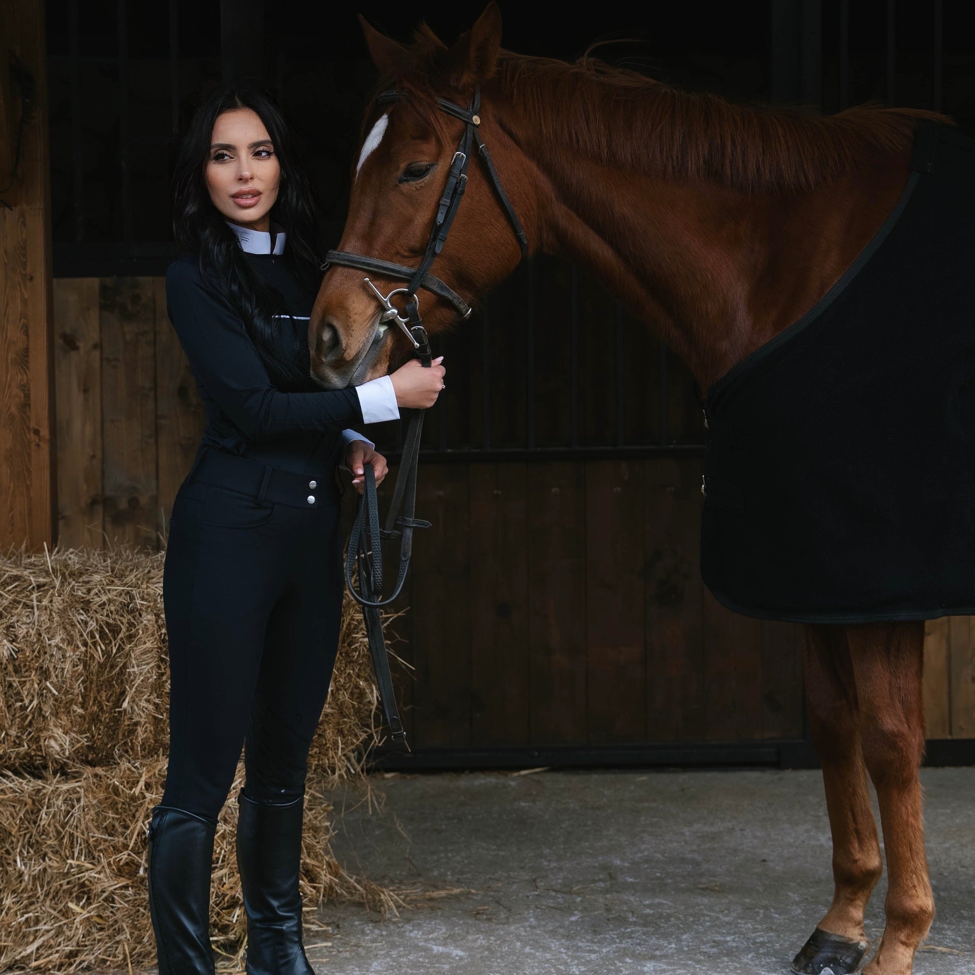 Woman in equestrian attire standing next to a horse in a stable.