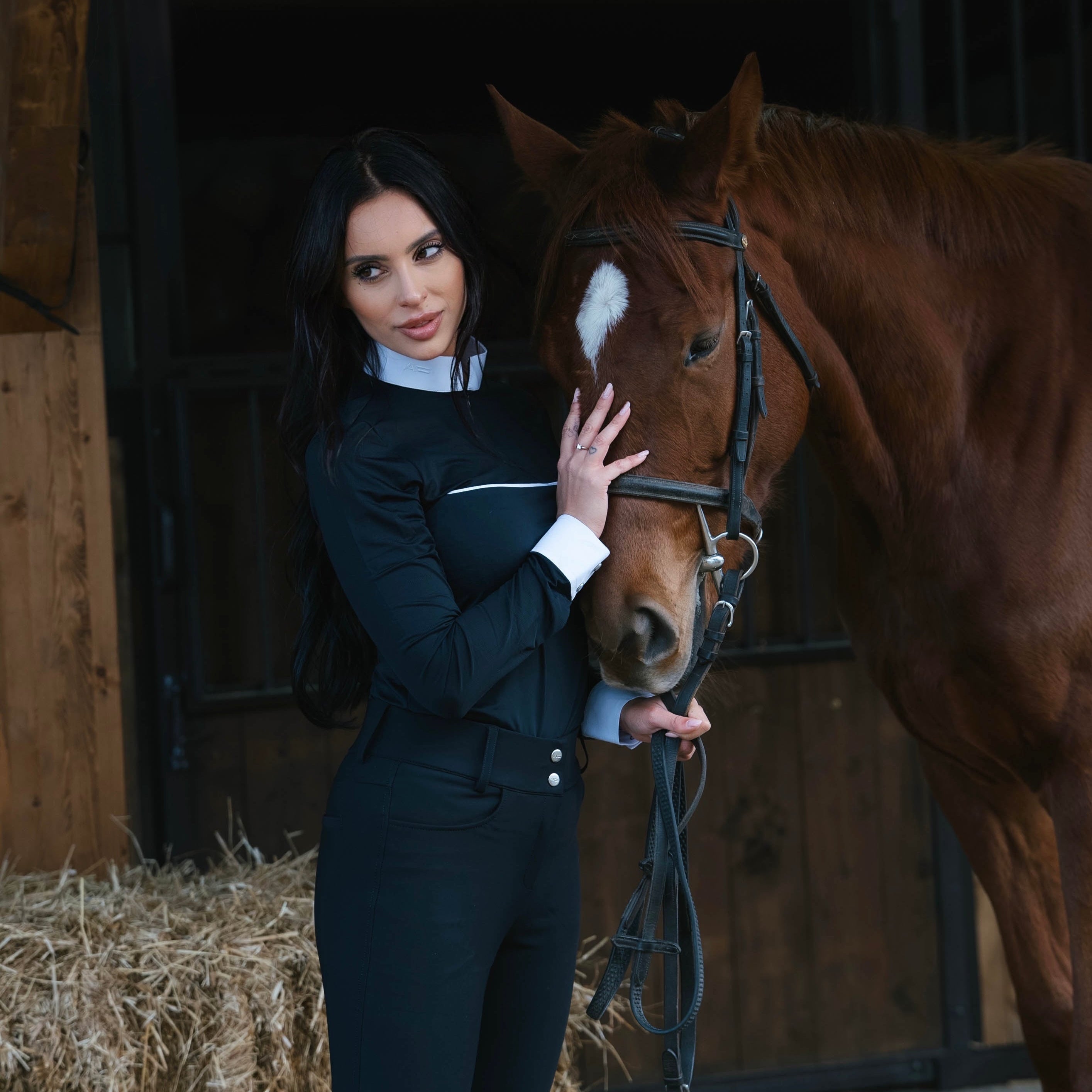 Woman in equestrian attire standing next to a horse in a stable.