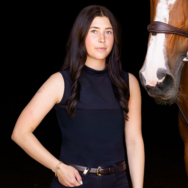 A woman in a black sleeveless top and black pants stands beside a brown horse with a white blaze and bridle against a dark background.